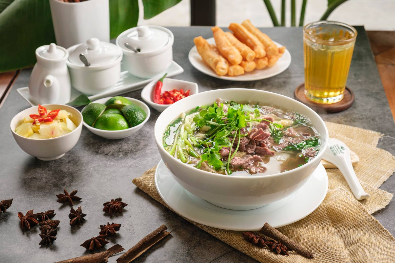 Pho bo with fresh herbs, lime, tea and fried dough sticks on a Hanoi breakfast table.