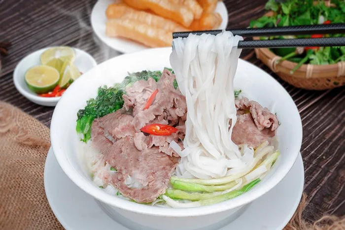 Pho bo noodles lifted with chopsticks from a steaming bowl beside herbs and fried dough.
